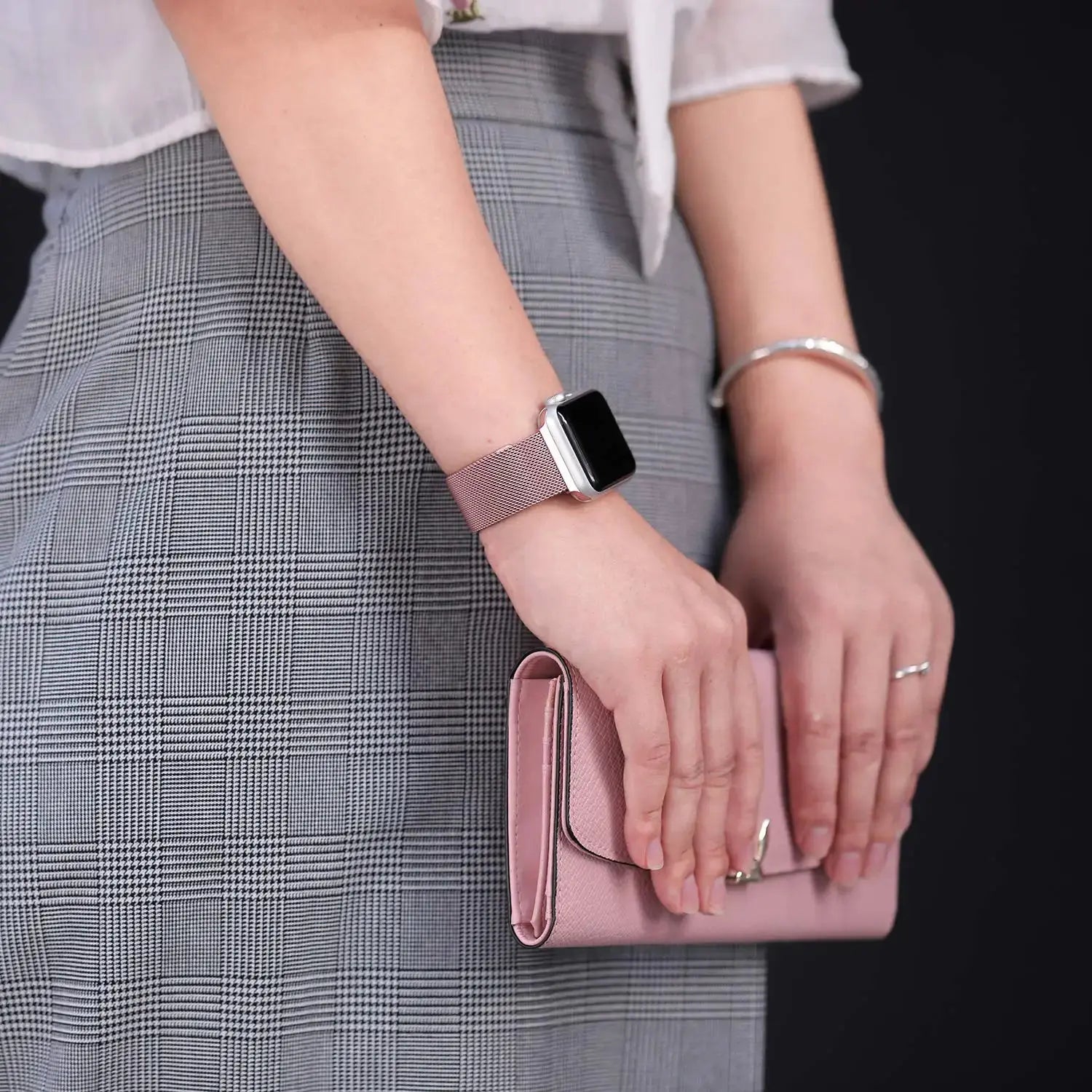 Woman wearing a Milanese strap with secure clasp on Apple Watch, holding a pink wallet, dressed in a stylish outfit.