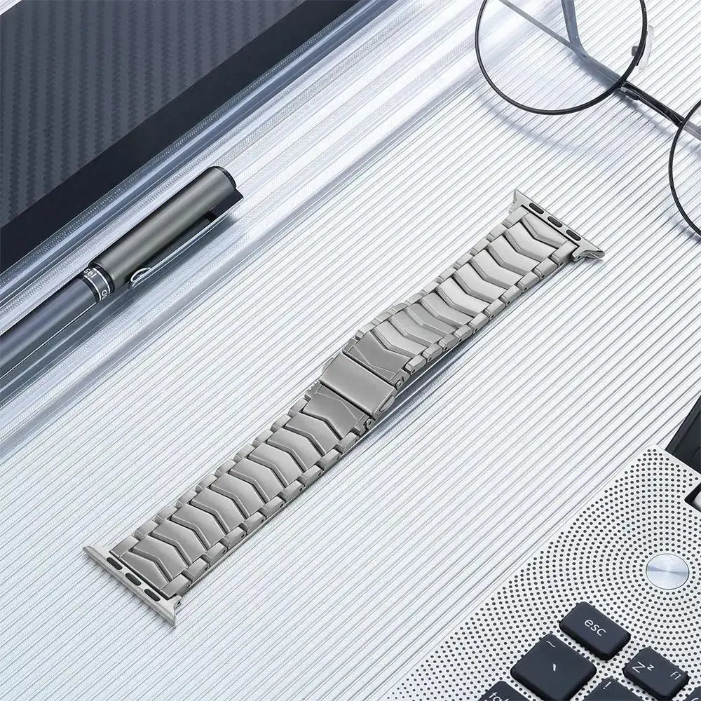 Grey satin-brushed titanium band for Apple Watch Ultra on a textured surface with a pen and glasses.
