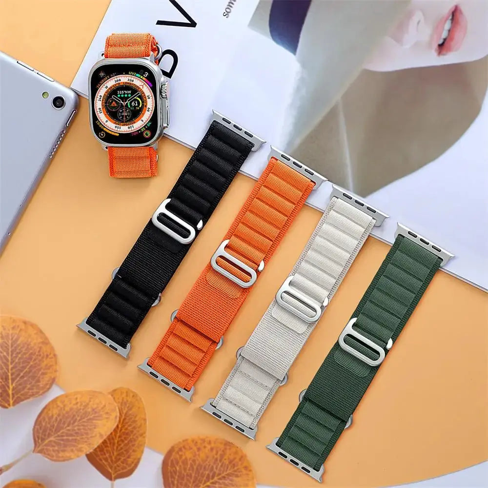 Rugged Alpine Loop watch bands in black, orange, beige, and green with stainless steel adjustment loops on an orange background.