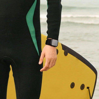 Man in wetsuit wearing Tactical Nylon Loop Bands for Apple Watch, holding a surfboard by the beach.