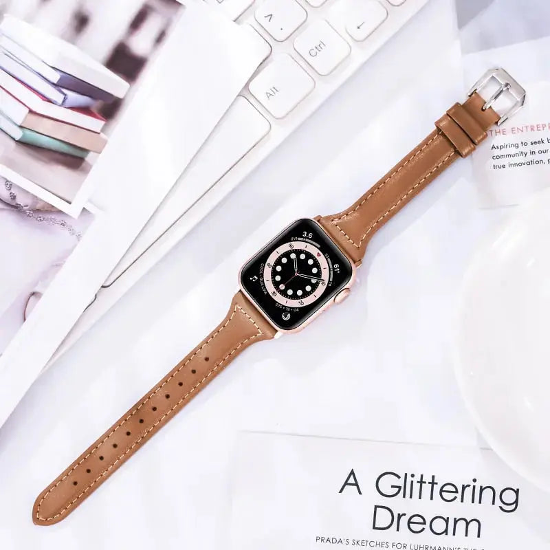 Slim leather band for Apple Watch, stylish tan strap with stainless steel connectors on a desk with notebooks.